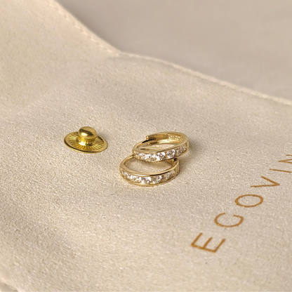 A pair of gold hoop earrings with a row of small, clear gemstones set around the circumference. The earrings are resting in a beige velvet pouch with the word "EGOVIN" embossed on it.