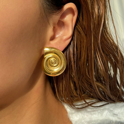 The image shows a women wearing gold-plated spiral statement earrings. The earrings are shaped like seashells and have a bold, eye-catching design. 