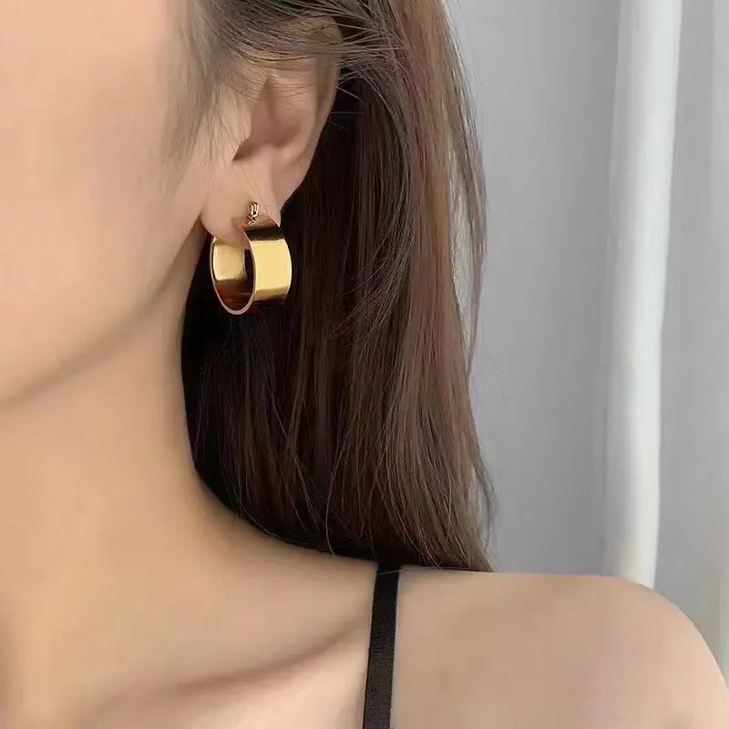 A close-up of a gold-plated hoop earring worn on a person's ear. The earring is large and flat, with a shiny metallic finish.