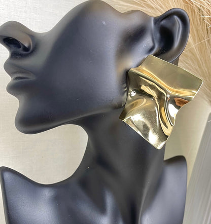 A pair of large, gold square earrings with a textured, crumpled surface. The earrings are worn by a mannequin with blonde hair.