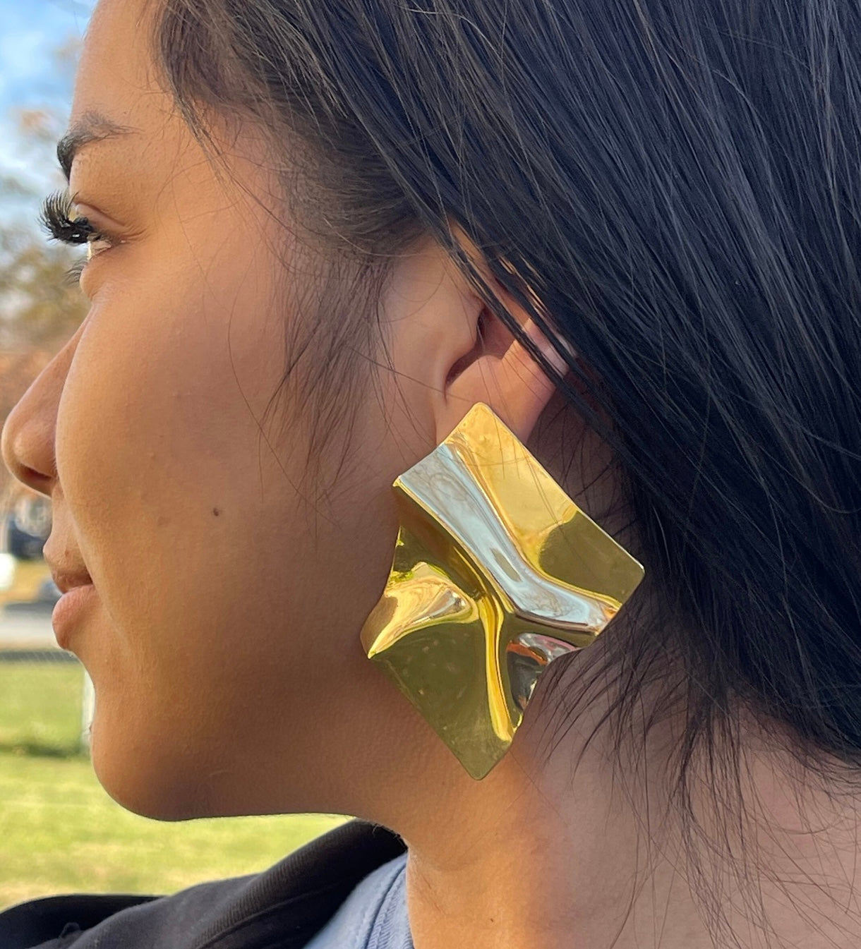 A large, gold square earring with a textured, crumpled surface. The earring is worn by a person with dark hair.