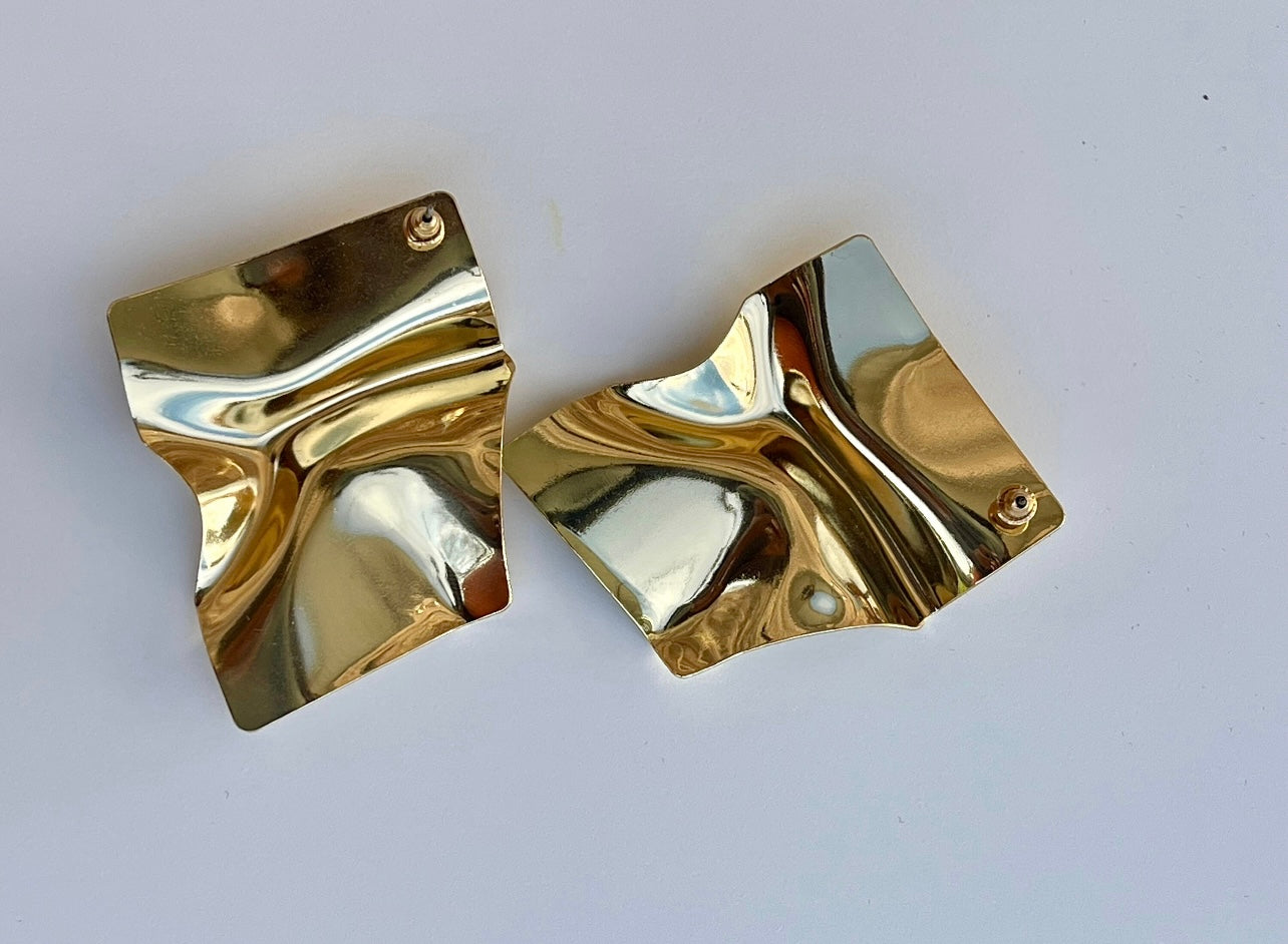 A pair of large, gold square earrings with a textured, crumpled surface. The earrings are resting on a white background.