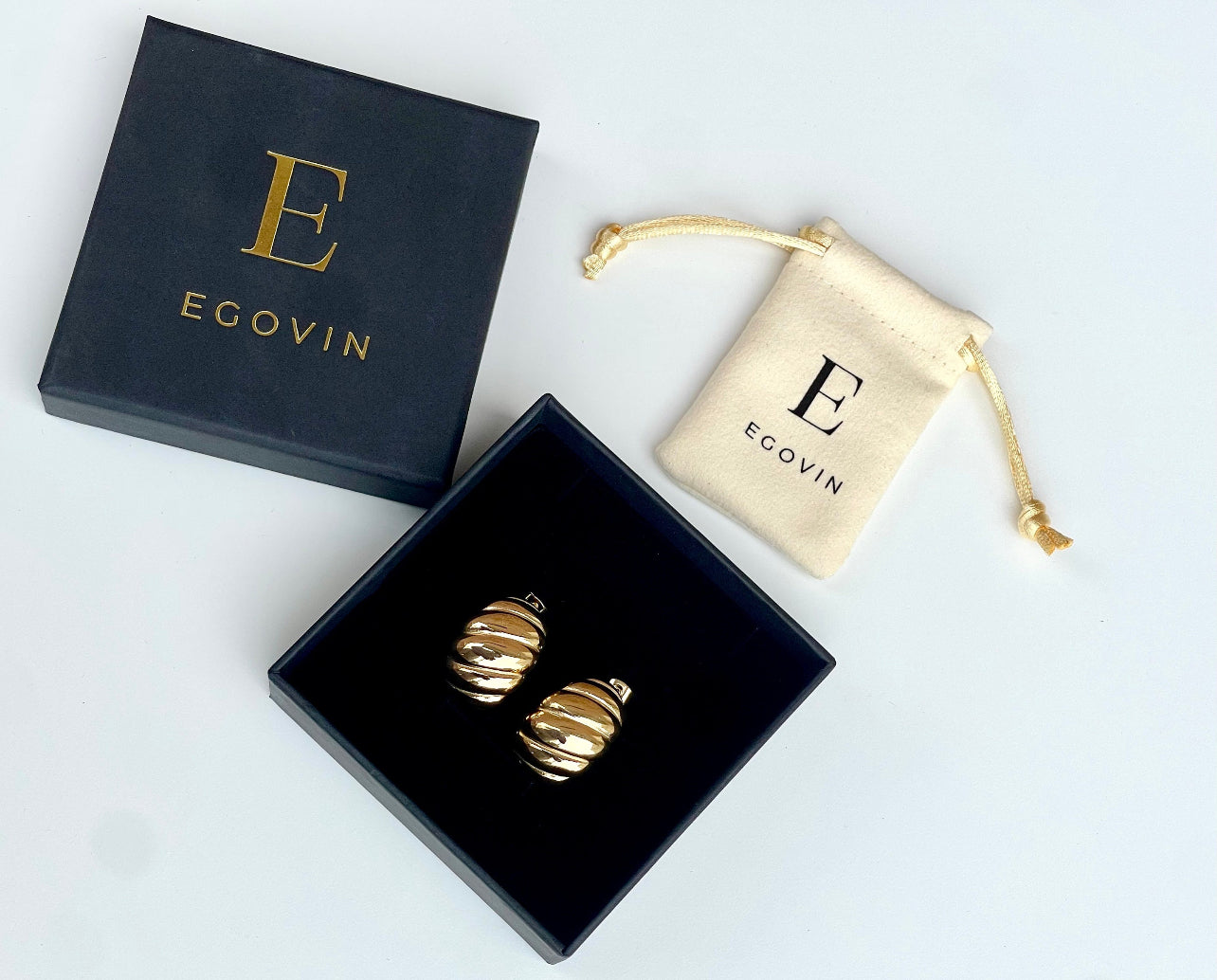 The image shows a pair of gold-plated textured chunky hoop earrings. The earrings come in a black gift box with a matching dust bag.  The earrings have a bold and modern design and are perfect for adding a touch of glamour to any outfit.