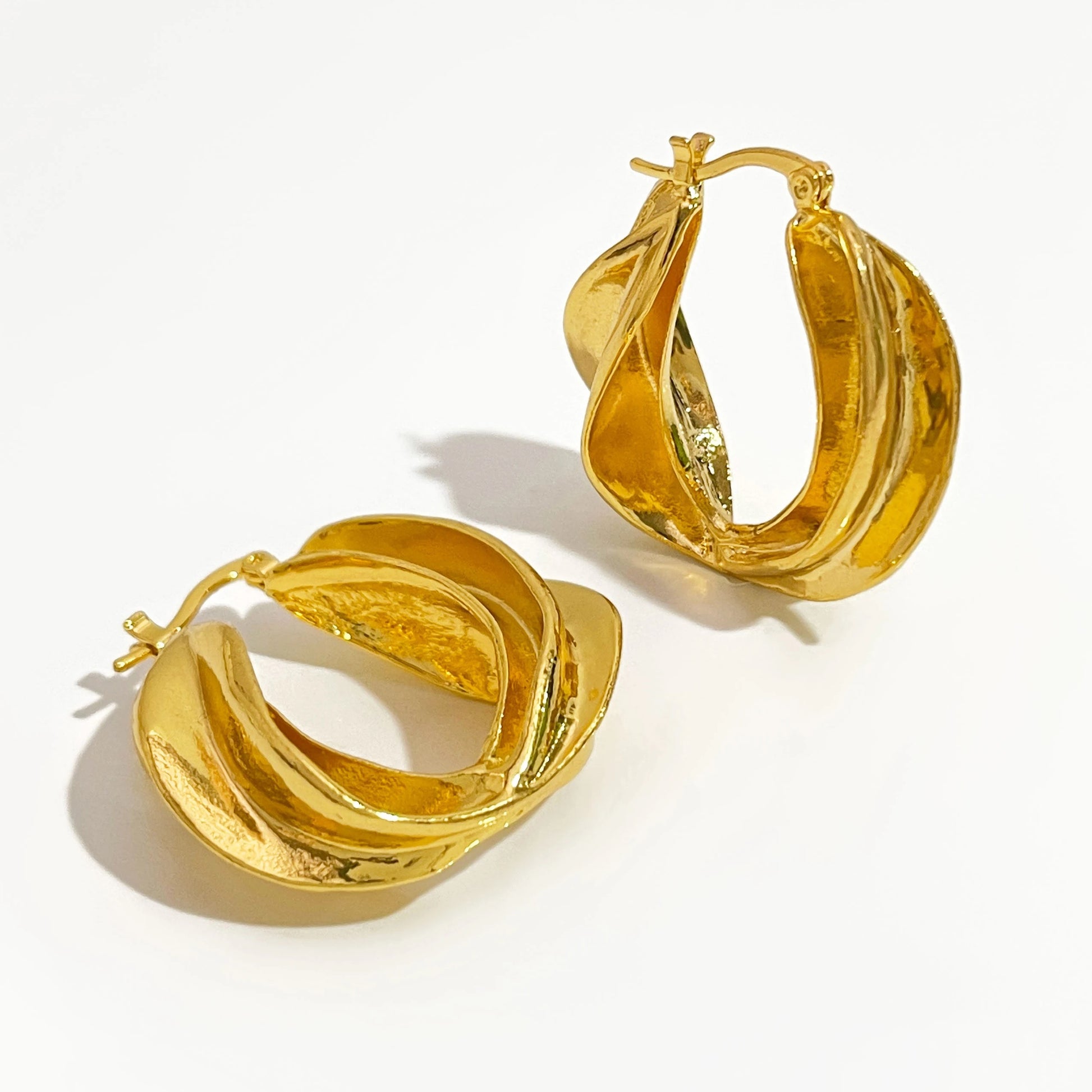 A pair of large, gold hoop earrings with a textured, wavy design. The earrings are lying flat on a white surface.