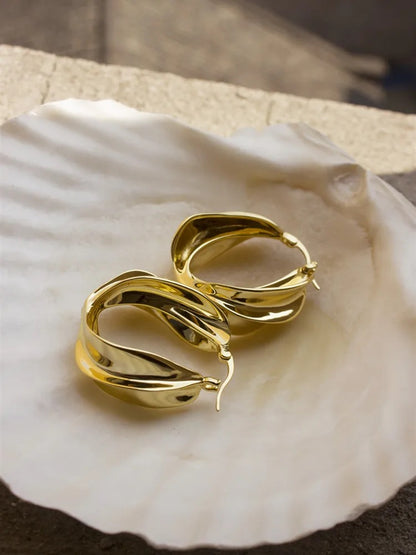 A pair of large, gold hoop earrings with a textured, wavy design. The earrings are resting on a white seashell.