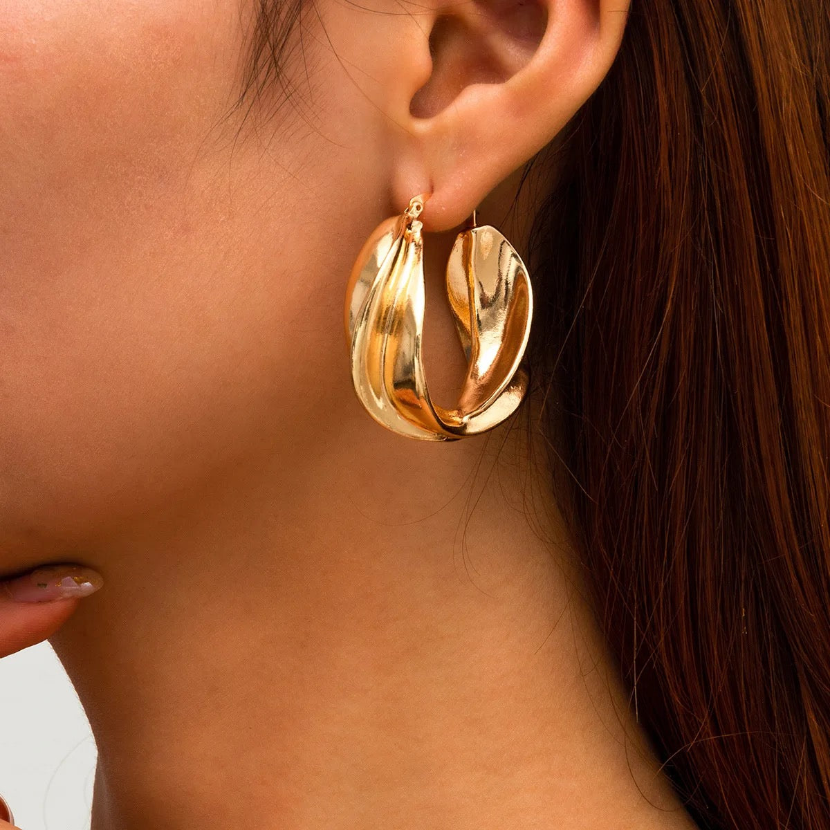 A pair of large, gold hoop earrings with a textured, wavy design. The earrings are worn in a woman's ear.