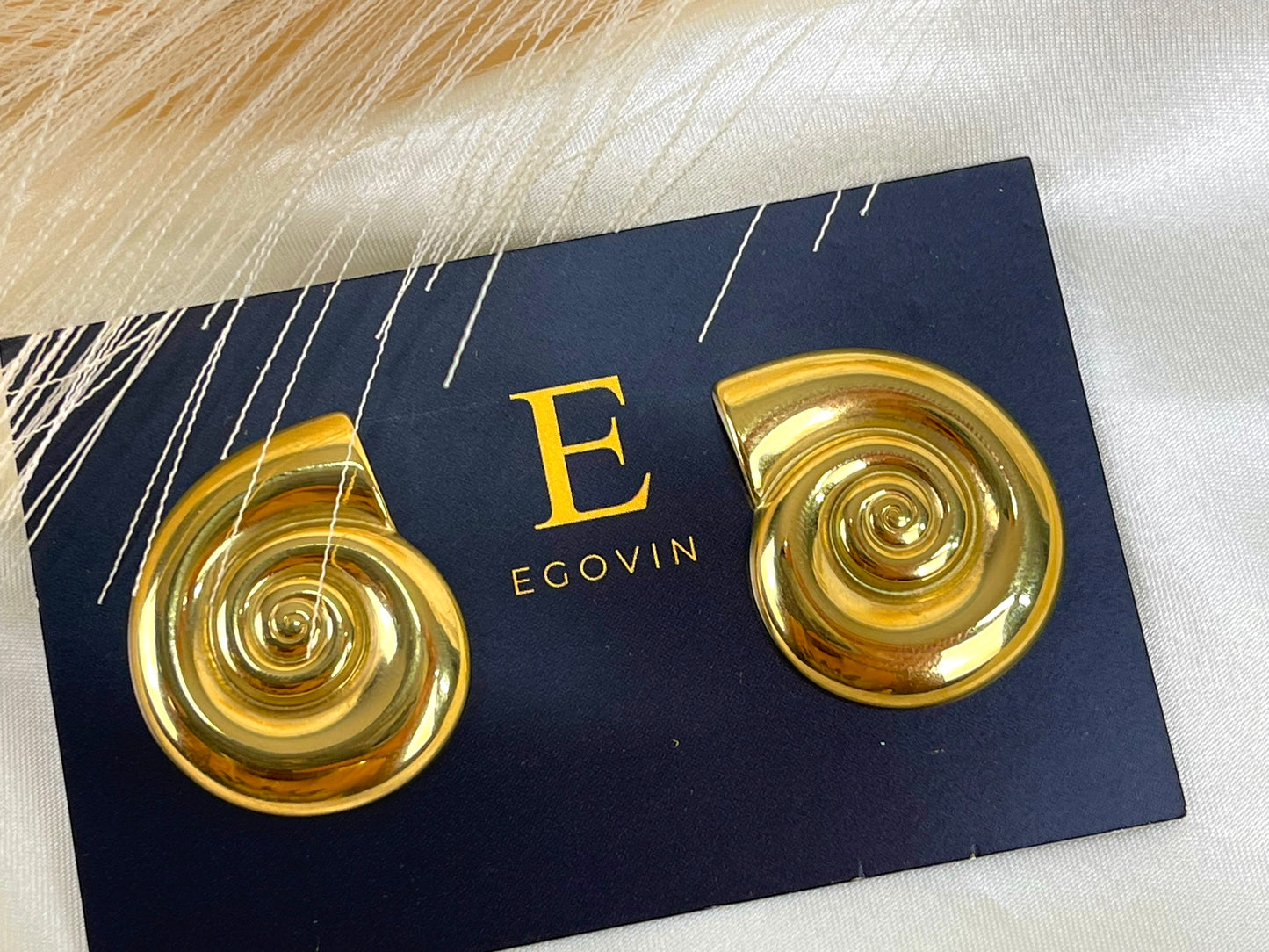 The image shows a pair of large, 18k gold plated spiral earrings with high quality material. They are shaped like seashells and have a glossy, metallic finish. The earrings are displayed on a card with the letter "E" and the word "EGOVIN" written on it.