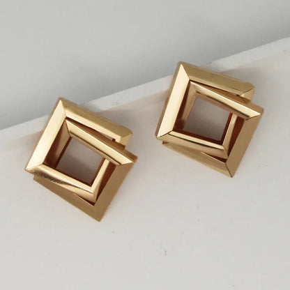 A pair of gold square earrings with a hollow center. The earrings are resting on a white surface.