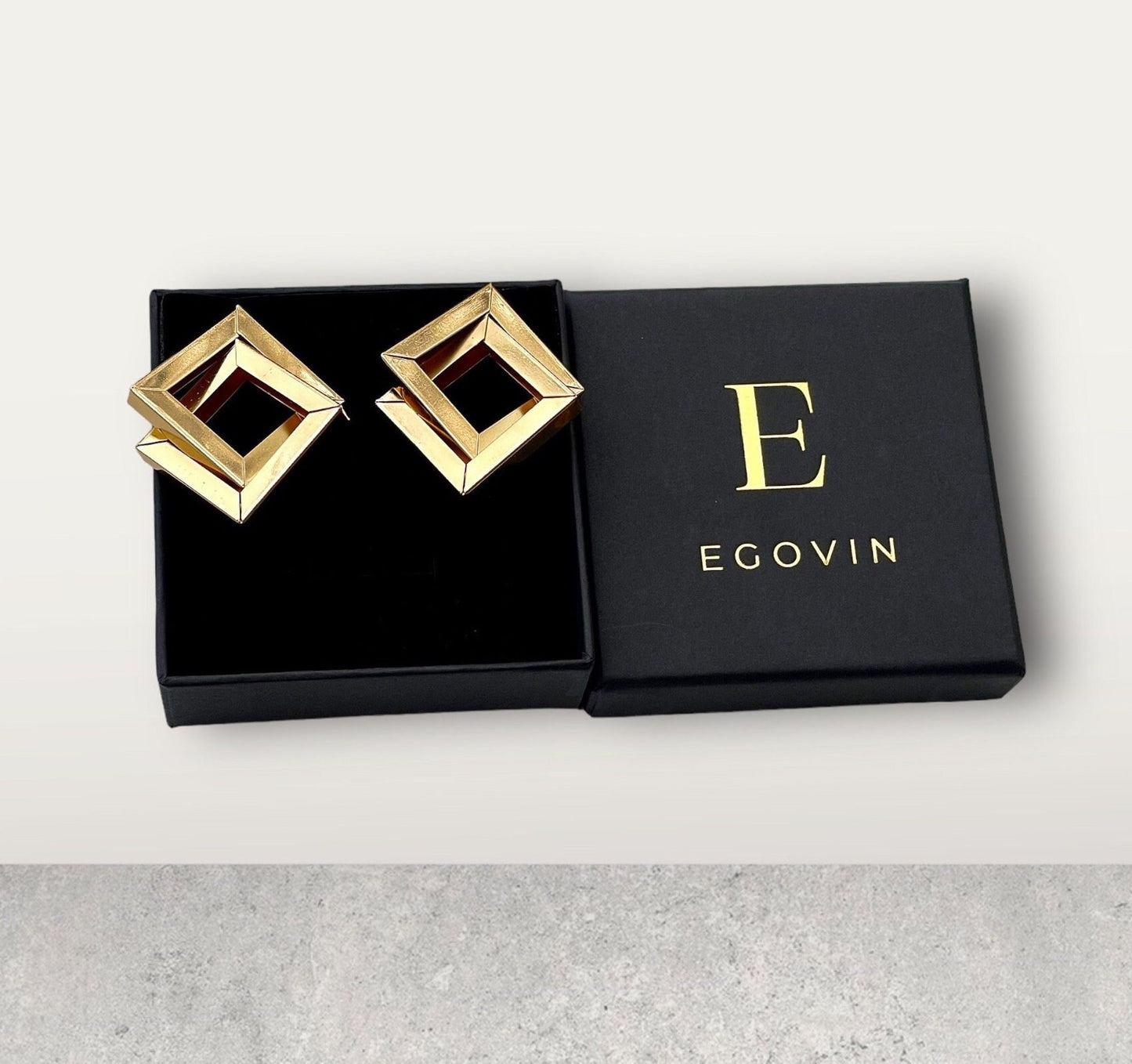 A pair of gold square earrings with a hollow center, resting in a black jewelry box. The box has the letter "E" and the word "EGOVIN" written on it.