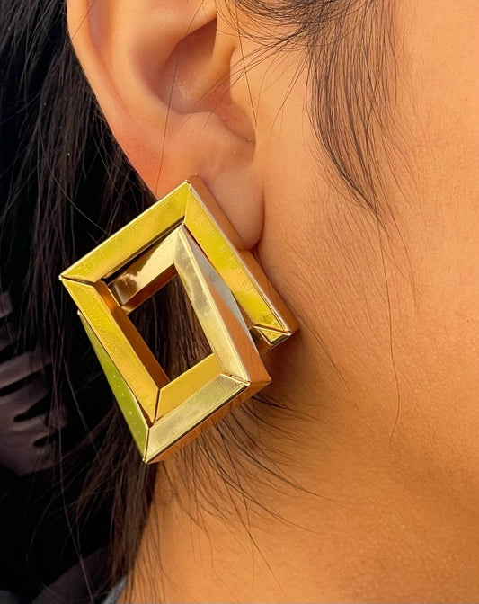 A pair of large, gold square earrings with a hollow center. The earrings are worn by a person with dark hair.