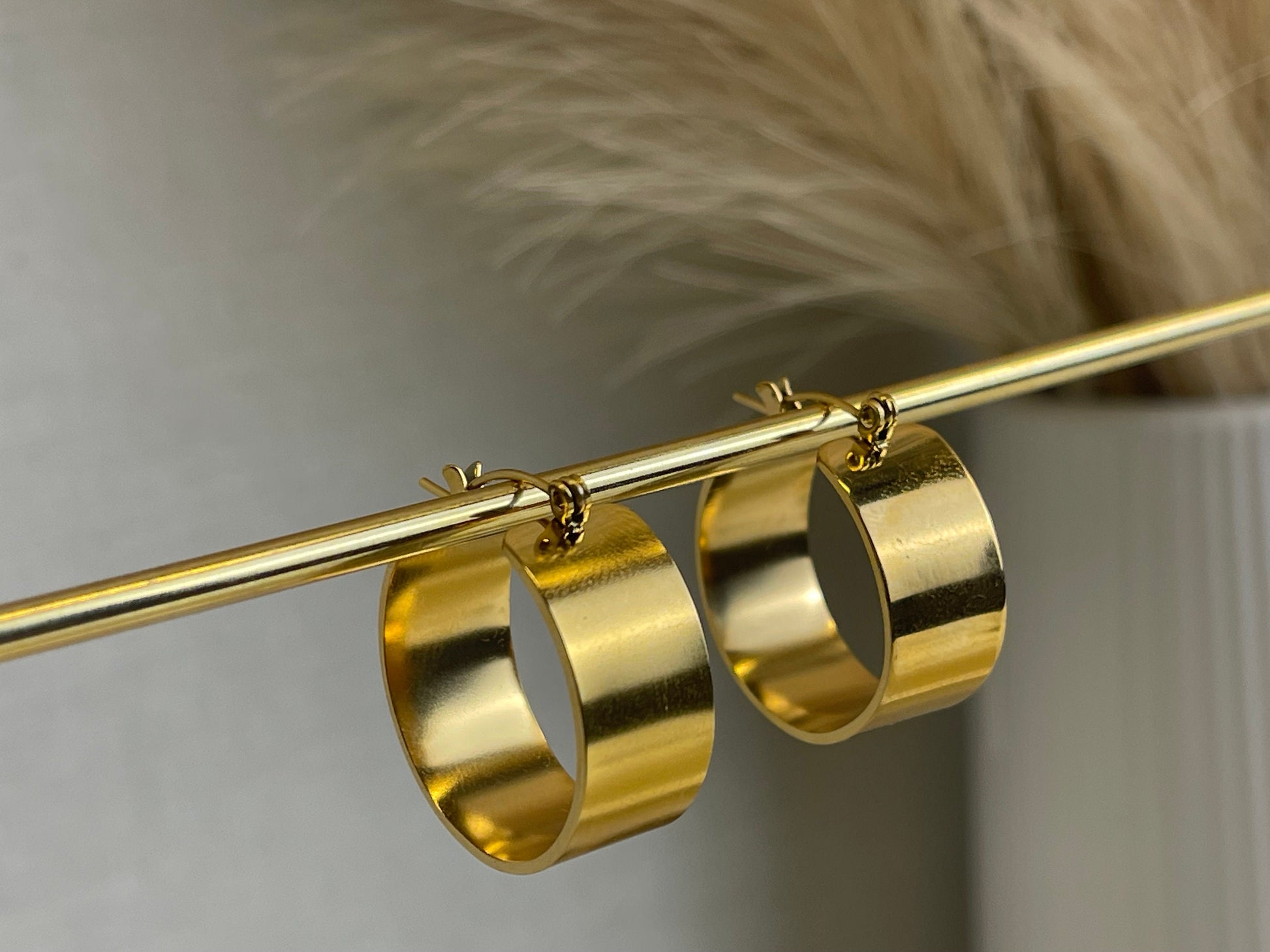 A pair of large, gold-plated hoop earrings with a flat, wide design displayed on a metal stand.